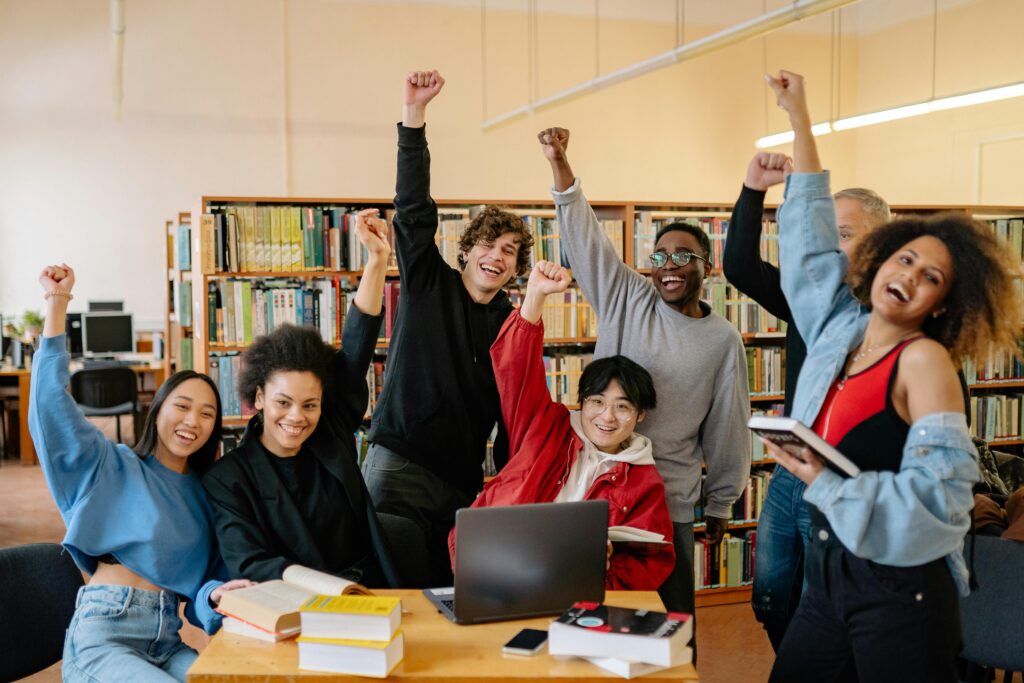 pexels-photo-8199562-8199562 Happy diverse students in library raising hands, enjoying academic success.